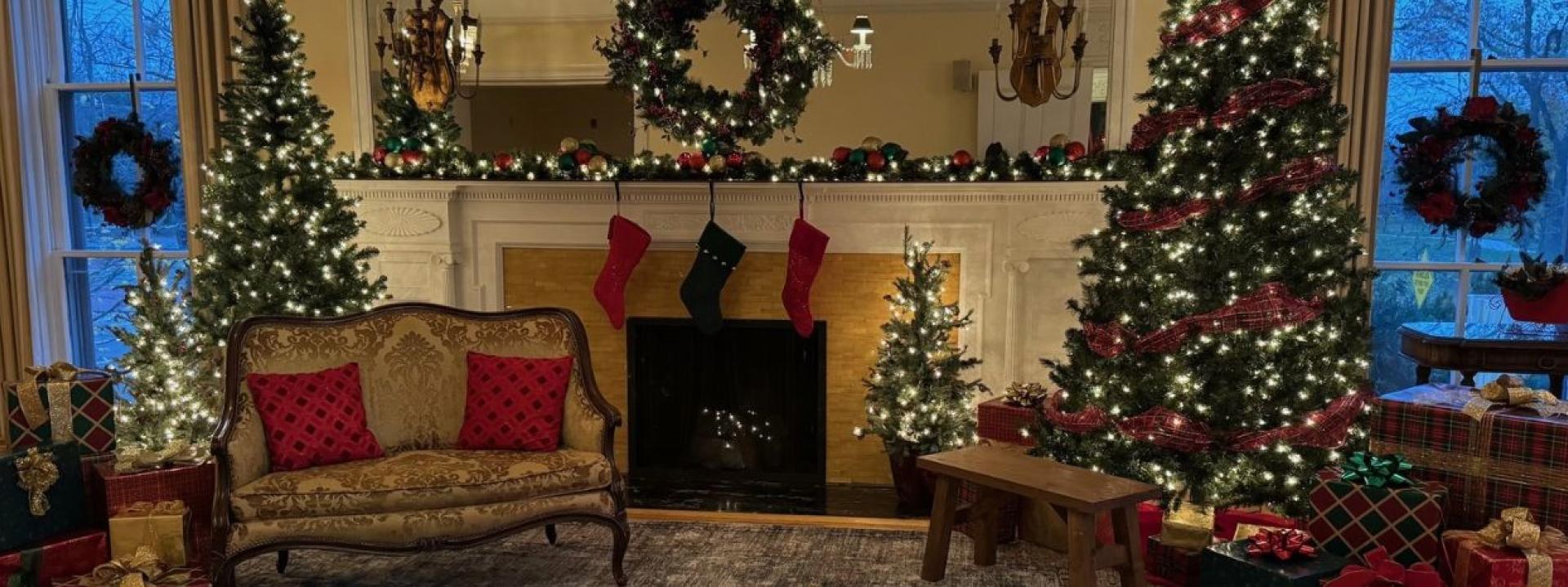 Cozy living room decorated for Christmas with a lit garland over the fireplace, a decorated tree by the window, wreaths, and a couch with red pillows, surrounded by wrapped gifts and twinkling lights.