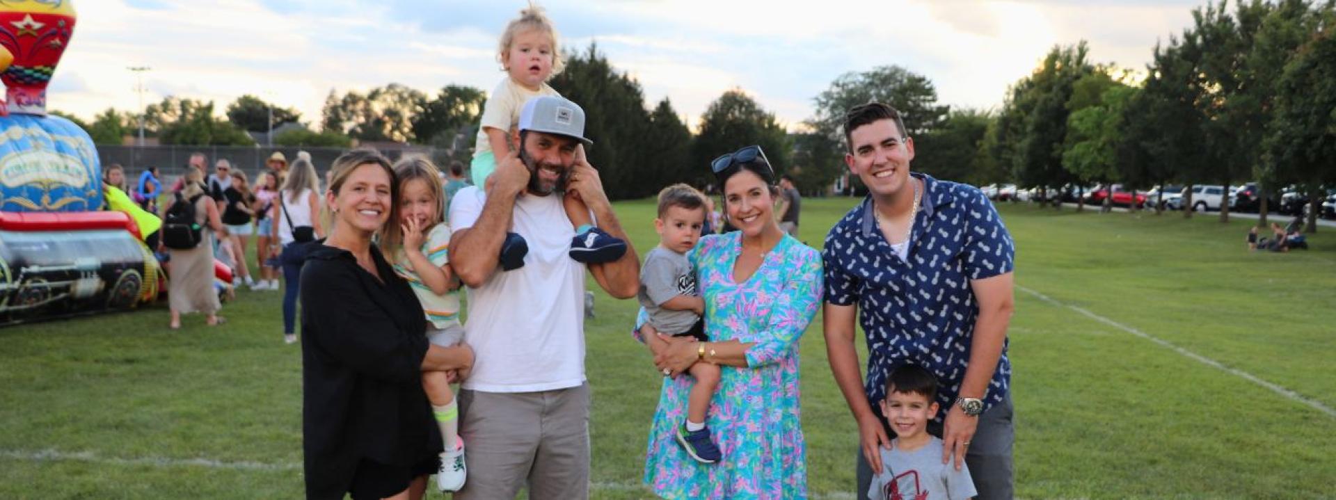 Family posing on a grassy field at an outdoor event, with a child being held up in the foreground and other kids nearby, while an inflatable play structure is visible at the left.