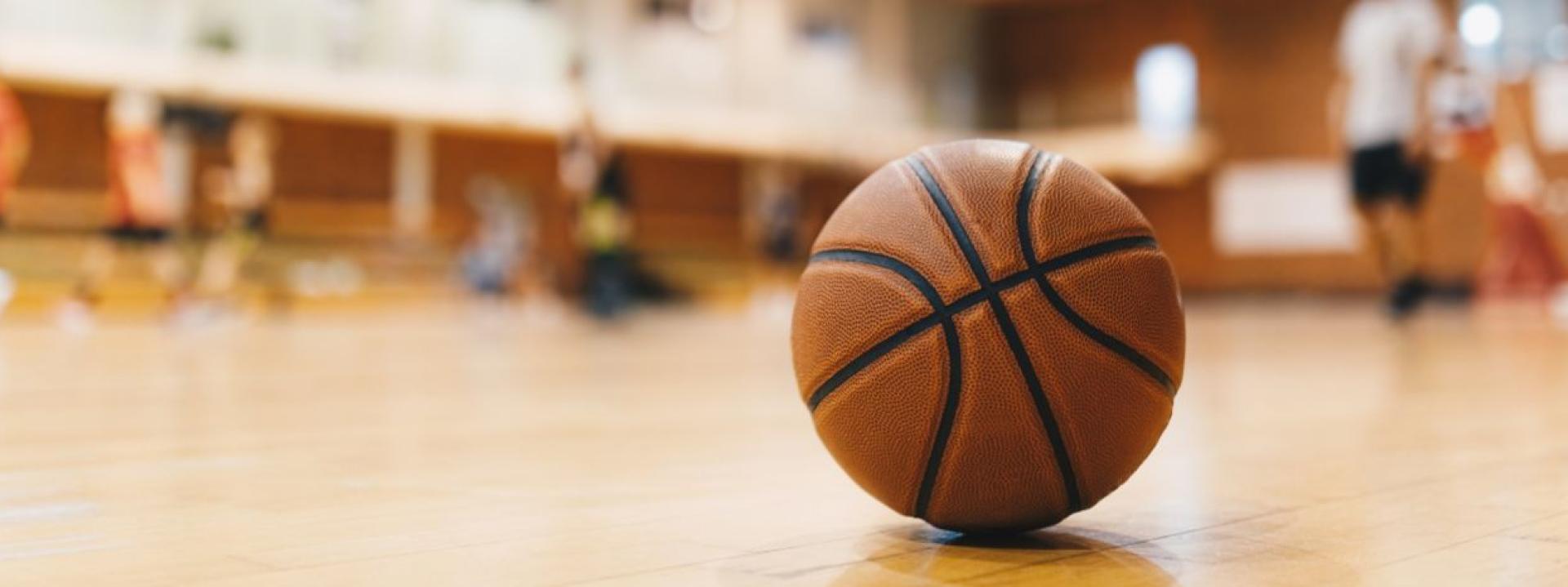 Basketball resting on a wooden gym floor, with a blurred indoor sports court and benches in the background.