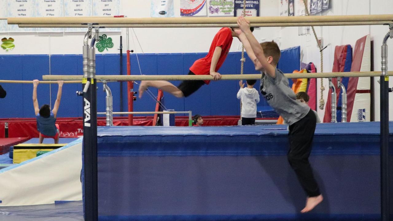 A gymnast reaches toward parallel bars with other gym equipment in the background.