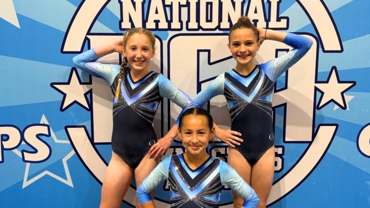 Three gymnastics in matching blue uniforms pose with hands on hips in front of a blue backdrop.