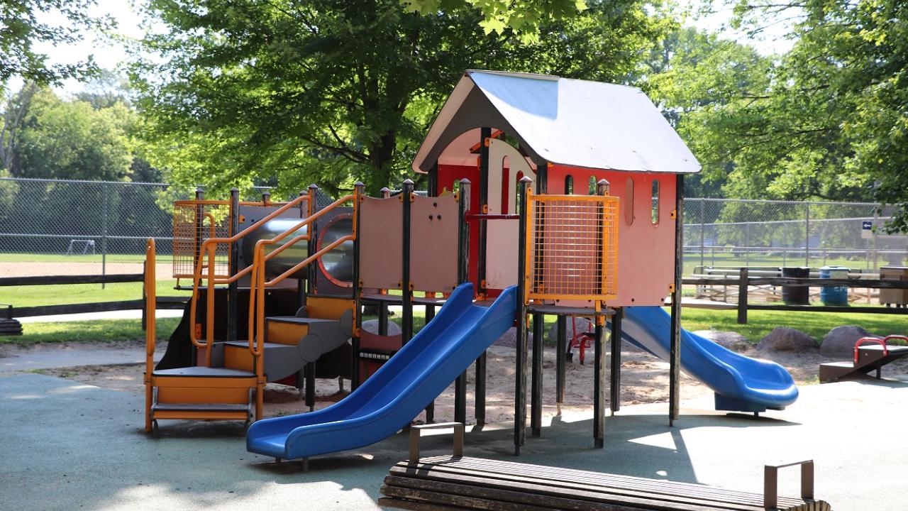 Playground equipment with two slides, stairs and a miniature house structure with a roof. In the background is a sports field.